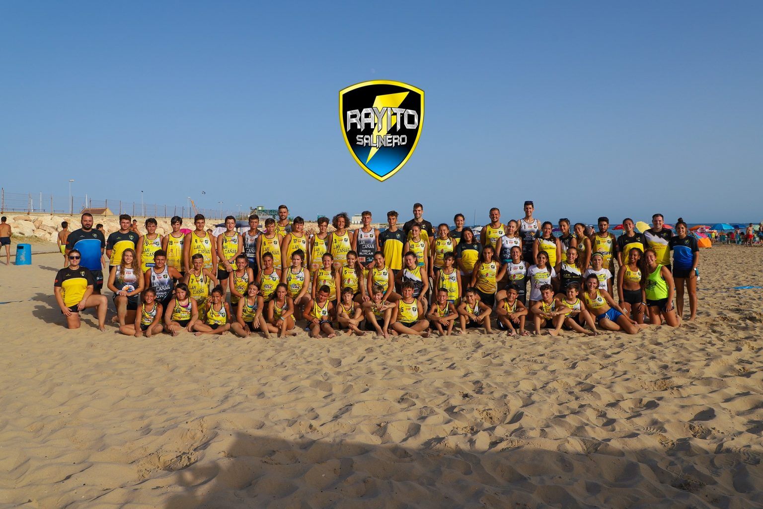 foto grupal de jugadores y entrenadores del CBMP Rayito Salinero en la playa con el escudo del club en el cielo